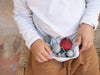 A child sits on a woven chair, using their white shirt as a pouch to hold colorful Grapat Ornament Set - Wish Balls, while wearing brown pants.