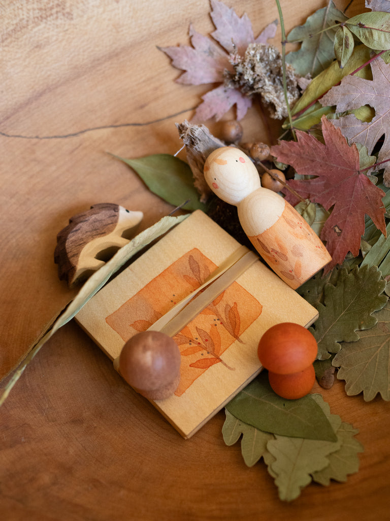The Hello Autumn Set (Grapat X Ostheimer Limited Edition) features wooden toys—a hedgehog, pegged doll, and orange balls—beautifully arranged with a decorated box and autumn leaves for an autumn-themed display.