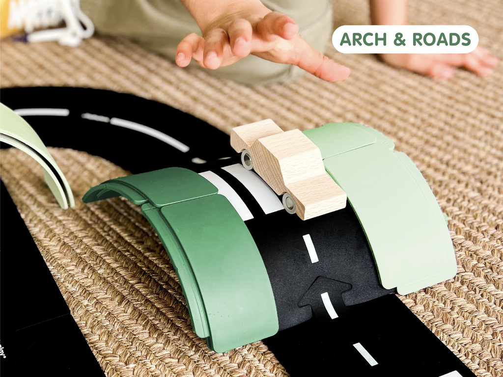 A child’s hand reaches toward a sustainable toy wooden car on a black road track with green WaytoPlay: Arches - Builder Set pieces arranged on a woven rug. The text “ARCH & ROADS” appears in the top right corner.