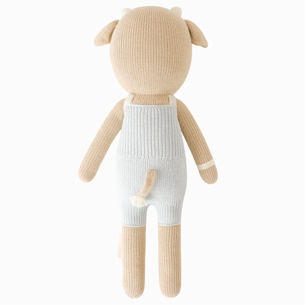 The Cuddle + Kind Asher The Cow is a hand-knit cotton plush toy shaped like a light brown cow, shown from the back in light blue overalls. It features small horns, a tail, hypoallergenic polyfill, and no visible face in this view.