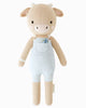 Cuddle + Kind Asher The Cow is a beige, hand-knit cotton stuffed cow with small horns, embroidered eyes, and a pink nose. Dressed in light blue overalls with a blue flower on the pocket and filled with hypoallergenic polyfill.