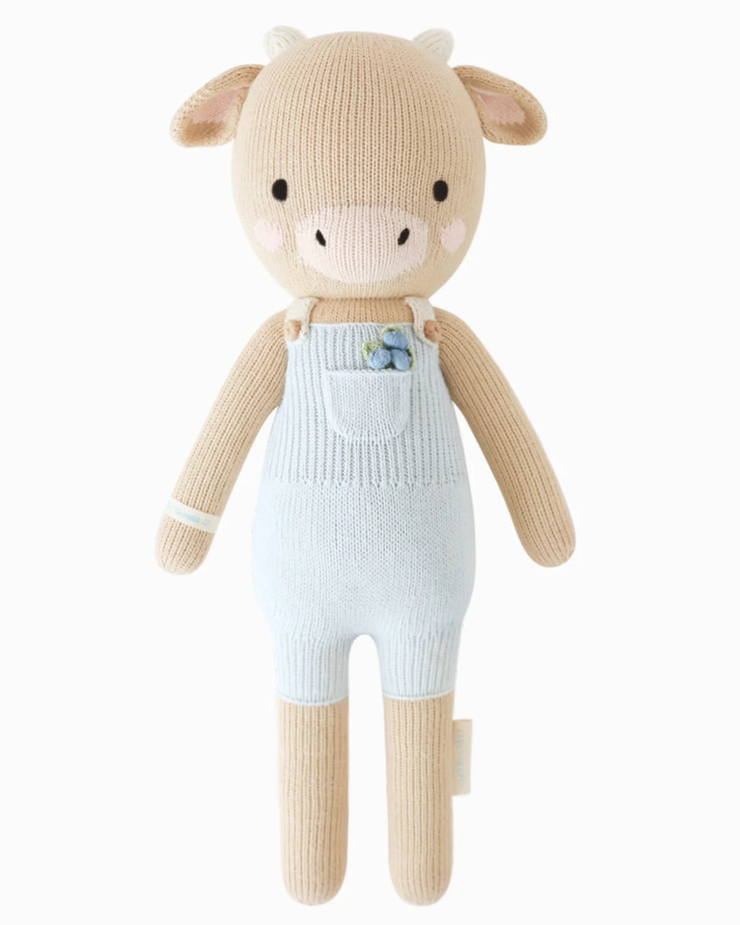 Cuddle + Kind Asher The Cow is a beige, hand-knit cotton stuffed cow with small horns, embroidered eyes, and a pink nose. Dressed in light blue overalls with a blue flower on the pocket and filled with hypoallergenic polyfill.