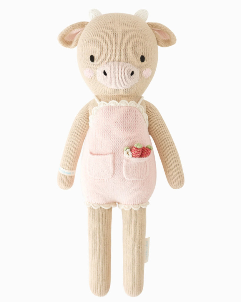 Cuddle + Kind Ava the Cow - Powder Pink is a hand-knit beige cow doll made from premium cotton yarn, wearing a pink apron with strawberry accents, and stands upright against a white background.