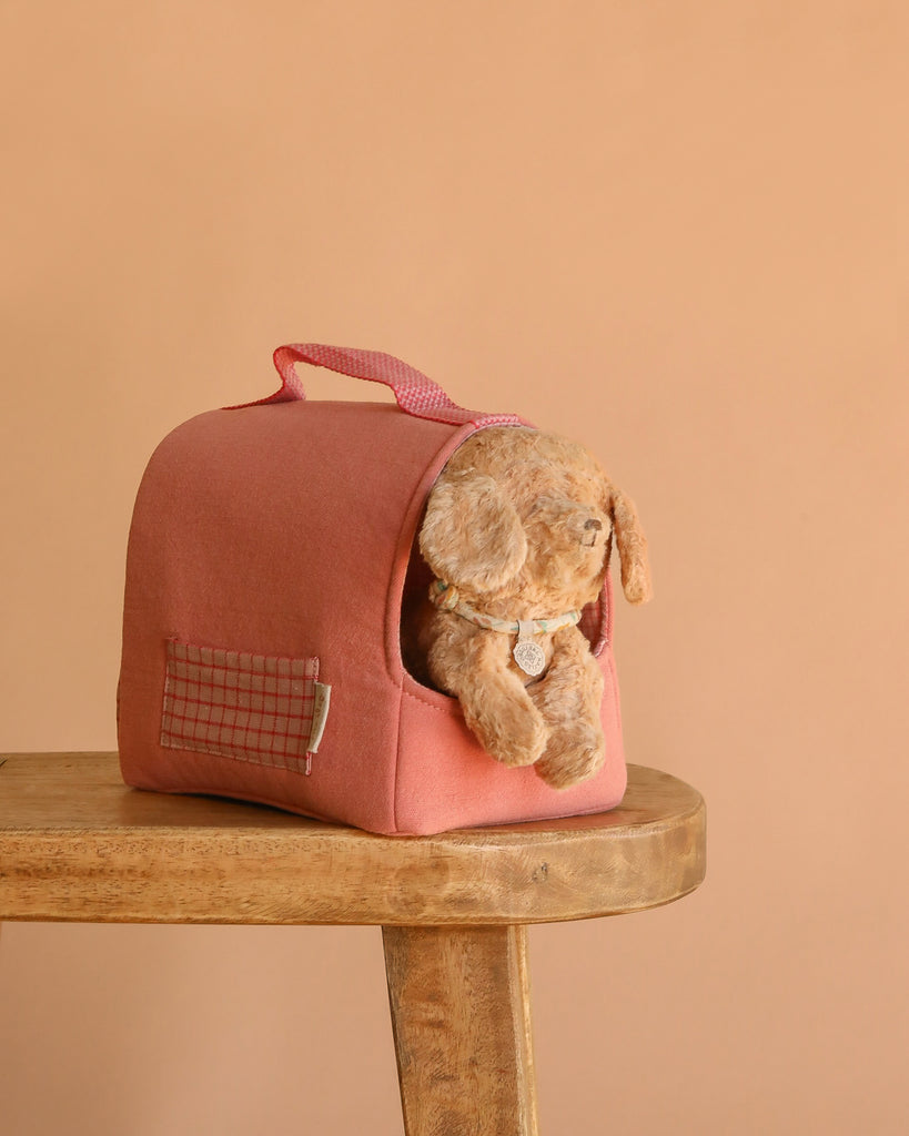 The Maileg Puppy in Coral Pet Carrier - Dusty Powder sits on a wooden stool. The soft pink carrier with a handle holds the plush puppy, set against a plain beige background.
