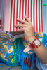 A person in a colorful embroidered outfit and a Mini Kyomo Watch - Cinnamon Roll holds a red-and-white striped book that covers their face.