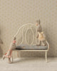 Three fabric bunny dolls in pastel dresses pose with the Maileg Vintage Bench, Miniature - Off White: one stands, one sits comfortably on the bench, and another leans playfully with its arms on it.