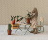 A stuffed bunny in a floral dress stands by a table potting a fabric plant. More greenery fills a crate on a bench, and the Maileg Flower Pot With Cactus, Miniature sits on the floor amid vintage-style décor.