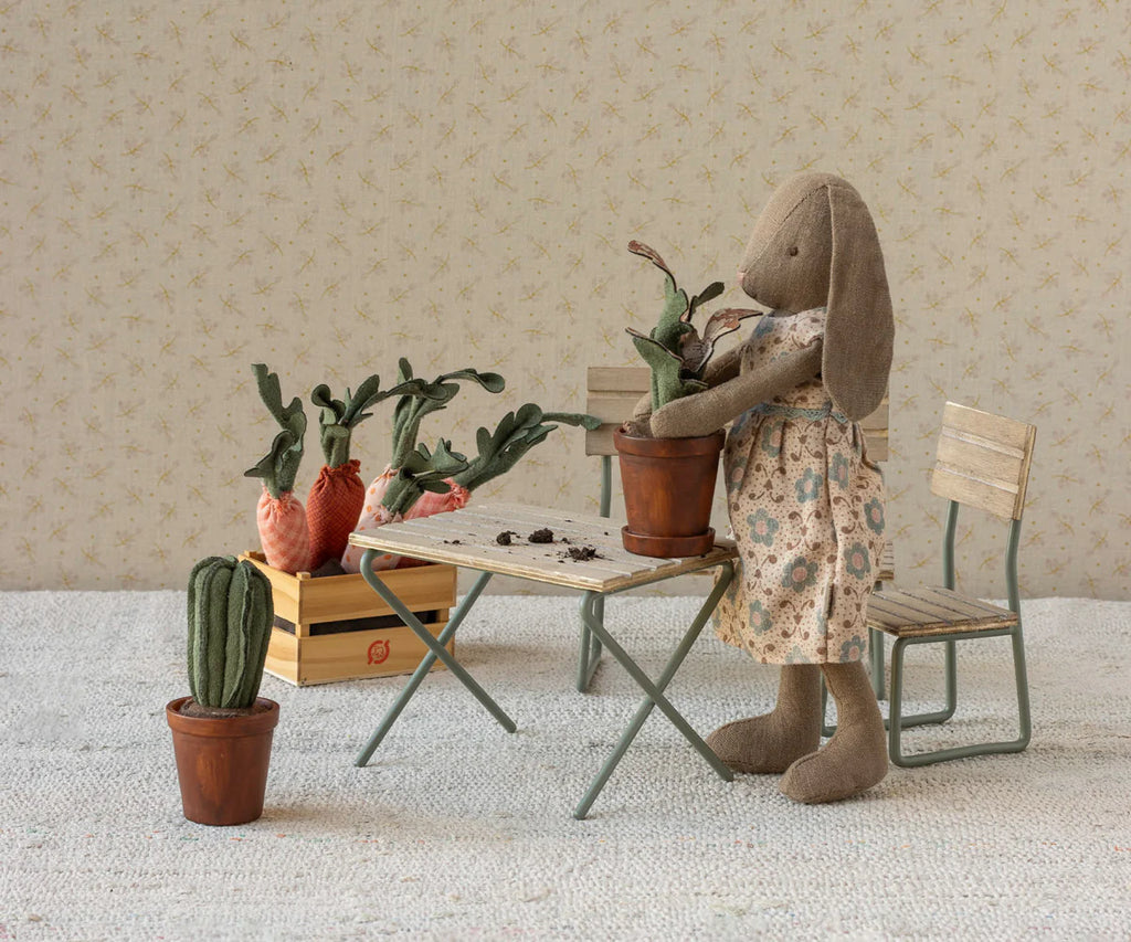 A stuffed rabbit doll in a floral dress tends to the Maileg Flower Pot With Plant, Miniature, beside a small table. Fabric vegetables and plants fill the table, crate, and floor, with wooden chairs and patterned wallpaper behind.
