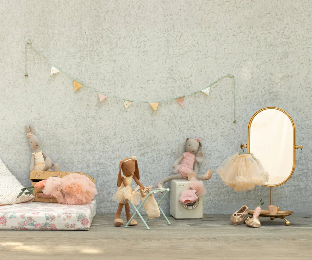 A pastel children's room features three Maileg Bunny, Size 2 - Classic - Ballet Suit and Skirt - Rose dolls, a gold-framed mirror, ballet slippers, and a garland on a light textured wall.