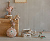 A woven basket with stuffed animals, a striped round pillow, a doll, unicorn-themed toys, and the Maileg Suitcase Set, 2-Pieces - Unicorn for toy storage are displayed on a light wooden floor against a gray wall with a framed picture above.