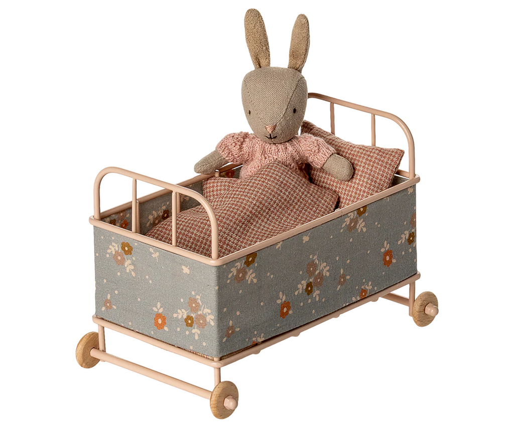 A PRE-ORDER: Cot Bed, Micro - Powder (ships early October) displays a stuffed rabbit in a pink dress, resting with a floral-patterned frame and pink-and-white checkered blanket and pillow.