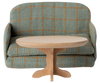 A small, oval wooden PRE-ORDER: Oval Coffee Table, Mouse - Nature (Estimated Ship Date: Early October) sits in front of a green plaid-patterned miniature sofa—perfectly mouse-sized, making for charming SuitCasa dollhouse furniture.