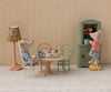 Three toy mice gather around the Maileg Oval Dining Table, Mouse Size - Nature, set with pastries and mugs, while one mouse opens a green cabinet. A small lamp glows nearby, creating a cozy dollhouse-like room.