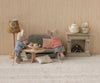 Two toy mice in striped shirts stand by the Maileg Oval Coffee Table, Mouse - Nature, with pastries. One mouse leans on the sofa, the other stands nearby. A lamp, fireplace, tea set, and cushion finish this cozy mouse-size SuitCasa scene.