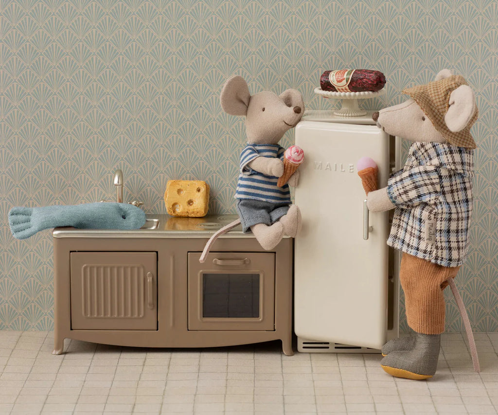 Two toy mice enjoy ice cream by the Maileg Fridge, Mouse - Off White in their kitchen, surrounded by dollhouse accessories. One sits on the counter while the other stands near a cheese wedge and fish toy, creating a cozy farmhouse scene.