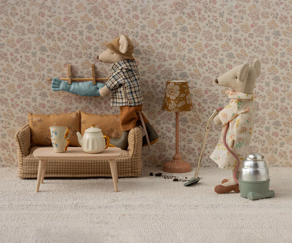 Two mice tidy their Mouse hole Farmhouse: one hangs laundry above the Maileg Couch, Mouse Size - Mable, while the other vacuums near a tea set in a vintage room with floral wallpaper and a lamp.