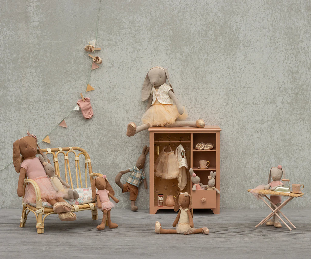 A group of stuffed bunnies in pastel clothes play in a cozy room featuring the Maileg Wardrobe, Large - Dark Powder, alongside a sofa, ironing board, and bunting against a neutral textured wall.