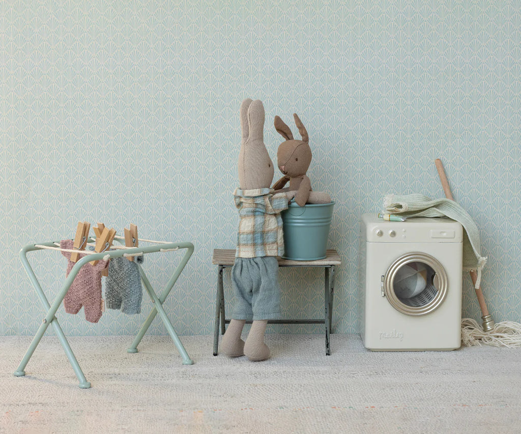 Two stuffed bunnies do laundry in a miniature scene with a washing machine, basket, and rack holding Maileg Extra Clothing: Knitted Suit, Micro - Heather for Micro Bunny. The larger bunny wears plaid shirt and blue pants, set against a pale blue patterned background.