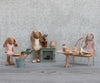 Three fabric bunny dolls in pink outfits sit in a cozy miniature home with a table, chairs, potted plants, and the Maileg Fireplace, Miniature - Dusty Green—all set against a gray textured wall background.