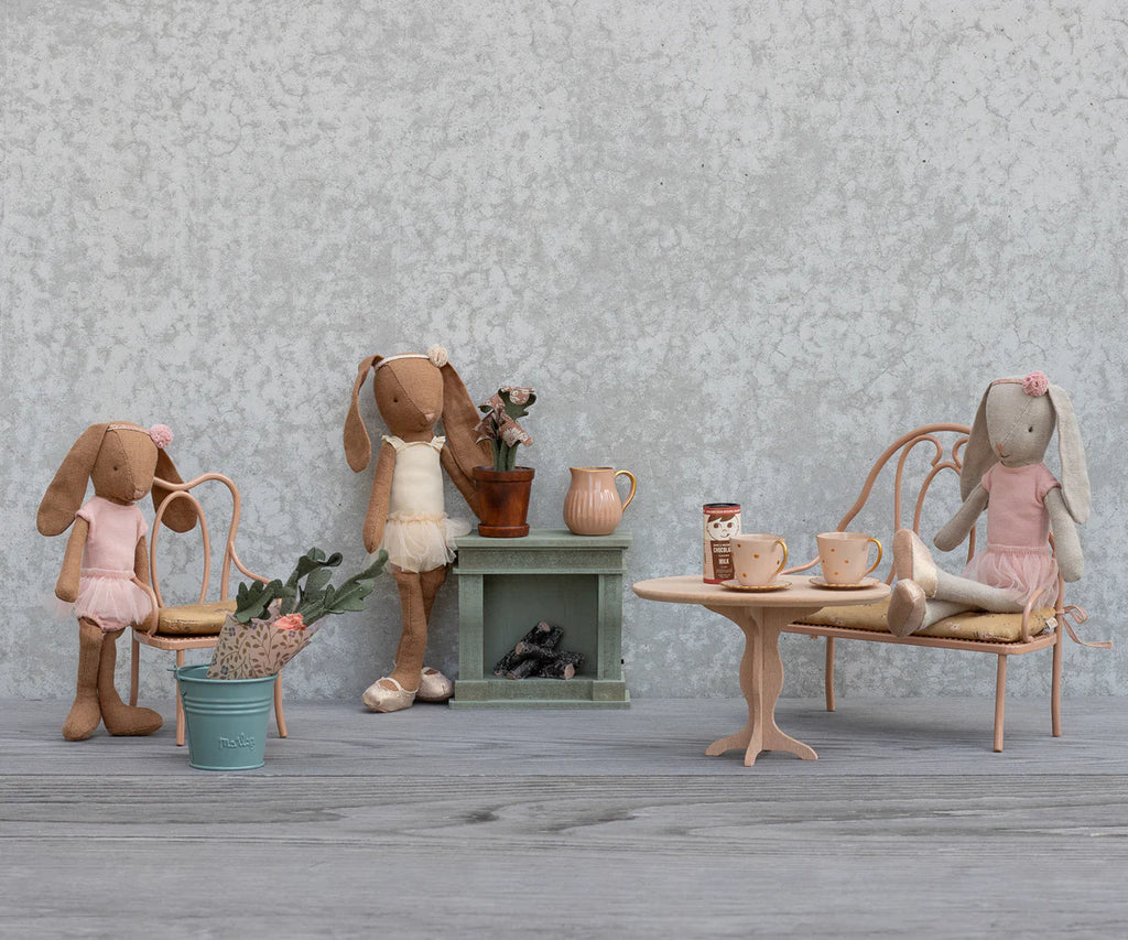 Three fabric bunny dolls in pink clothes have a mini tea party with Maileg’s Vintage Chair, Mini - Dark Powder, surrounded by tiny rabbit furniture, teacups, a plant, and a decorative fireplace against a textured gray wall.