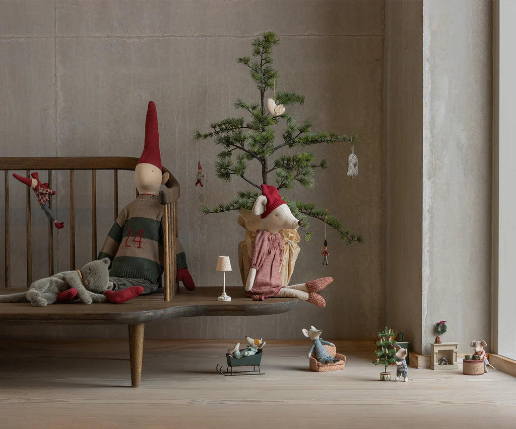 A cozy scene with Maileg Christmas Mouse, Maxi - Girl in festive clothes among other fabric dolls, a decorated tree, mini animal toys, and tiny furniture on a wooden bench and floor by a gray wall.