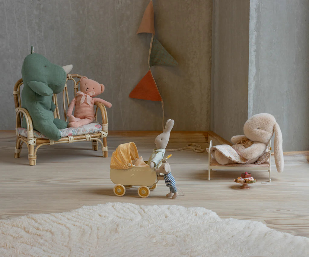 A Maileg Plush Bunny, Small - Latte lounges on a daybed while stuffed animals, including a green dinosaur and pink bear, sit nearby. A small gray bunny pushes a toy stroller. Colorful flag garland decorates the wall of the cozy room.