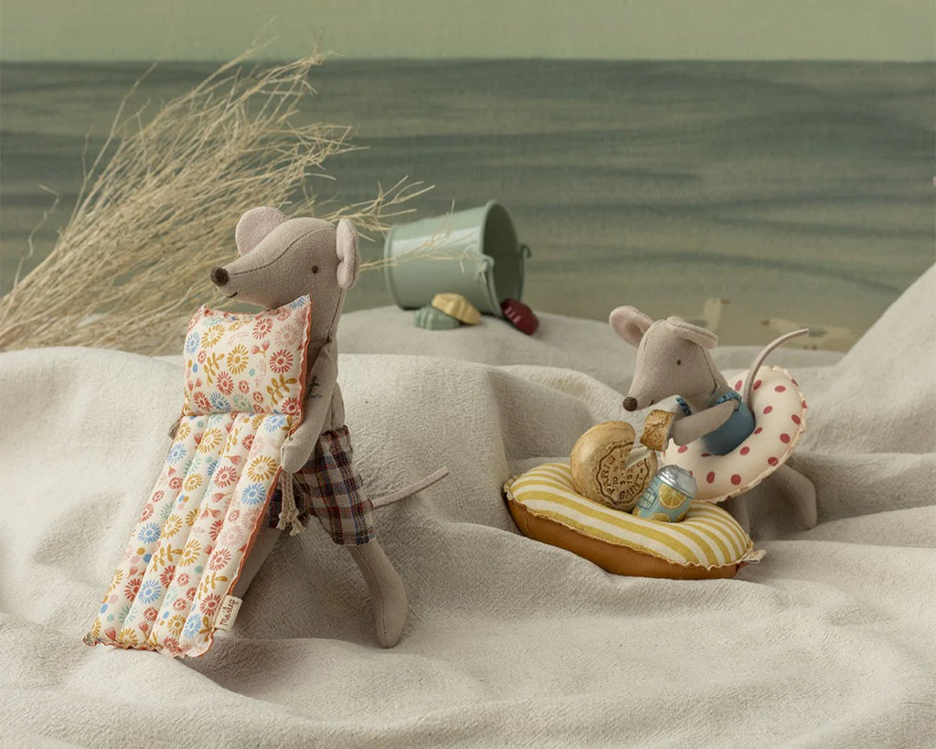 The Maileg Beach Mouse with Air Mattress - Dad features a fabric mouse in plaid shorts and floral float, plus an air mattress. Set against a beach scene with dried grass, a bucket, ocean waves, and other playful summer details.