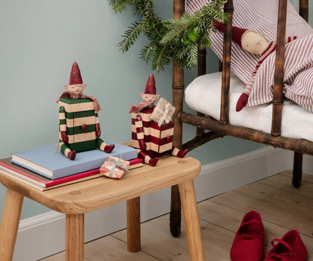 A PRE-ORDER Maileg Elf with Drawer - Red (Estimated Arrival Mid August) sits near books and a festive gift box, while a doll on a rustic chair and holiday décor like pine greenery and red shoes complete the charming scene.