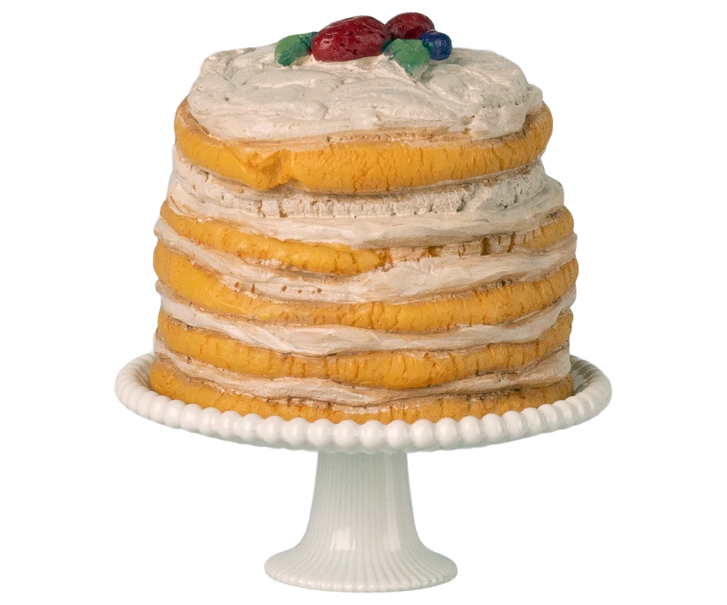 The Maileg Party Cake and Cake Stand, Mouse - Off White (Expected arrival: 05/01/26) features a tall layered yellow sponge cake with white frosting and red-blue fruit decorations, displayed on a fluted white stand as the party centerpiece.