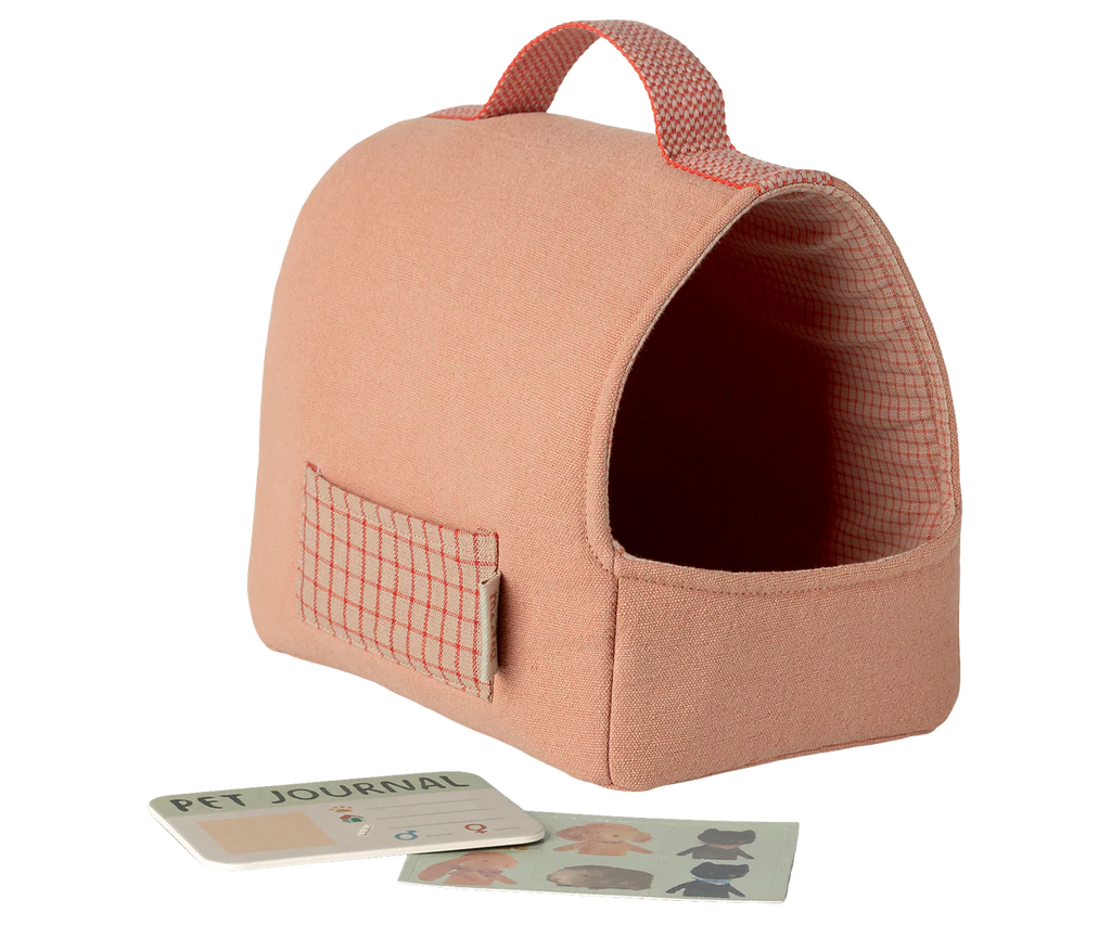 The Maileg Pet Carrier - Coral, a soft pink carrier with a handle and side pocket (expected by 02/15/26), sits beside a small pet journal and sticker sheet—stylish accessories arranged on a light background.