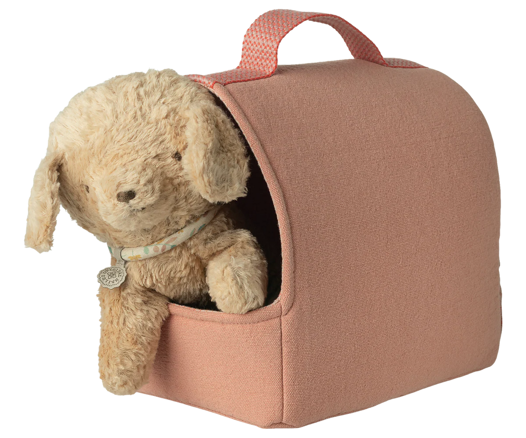 A soft puppy plush with a collar peeks out of the Maileg Pet Carrier - Coral, which has a handle on top and an open front. One paw and the pup’s head are visible against a plain white background.
