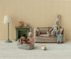 A cozy scene: an adult Maileg Micro Bunny With Ball - Brown - Dusty Rose (Magnetic Hands) sits on a sofa with a child bunny, another kneels by a baby in a basket. A lamp, fireplace, and the soft bunny toy ball add extra charm to the room.