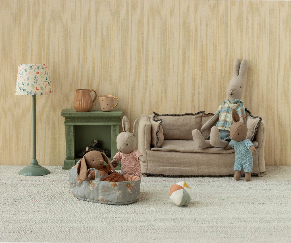 A cozy scene: an adult Maileg Micro Bunny With Ball - Brown - Dusty Rose (Magnetic Hands) sits on a sofa with a child bunny, another kneels by a baby in a basket. A lamp, fireplace, and the soft bunny toy ball add extra charm to the room.