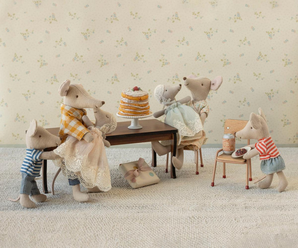 Six Maileg mice from the adorable mice family gather around a table with the Maileg Party Cake and Cake Stand, Mouse - Off White, as one mouse stands on a chair pouring syrup. A wrapped gift rests beneath the table, while all the mice are dressed in cute, colorful outfits.