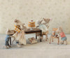 Five plush mice gather around the Maileg Long Dining Table, Mouse, which is topped with a tall layered cake. One mouse stands on a chair, preparing tea, while a wrapped gift sits on the floor. The scene feels cozy and whimsical, enhanced by charming dollhouse accessories and soft pastel tones.