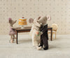 Two toy mice from the Maileg mice family, dressed as a bride and groom, dance together, while another mouse in a purple dress sits at a table featuring the Maileg Party Cake and Cake Stand, Mouse - Off White. A gold chair rests beside the table on a textured surface.