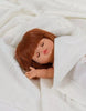 A small Minikane Baby Doll With Sleeping Eyes (13") - Capucine with straight brown hair and closed eyes is wrapped snugly in a white knitted blanket. The doll has a peaceful expression, its right hand slightly raised, and exudes a natural vanilla scent against a soft, knitted white fabric background.