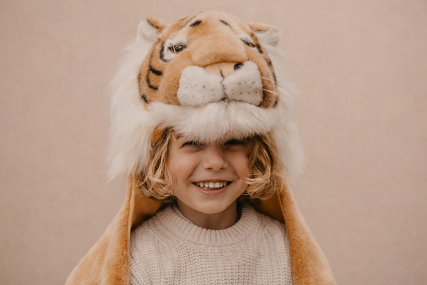 A smiling blonde-haired child wears the Costume - Tiger, a large plush faux fur hat with ears and face, paired with a cream knit sweater, standing against a plain beige background.