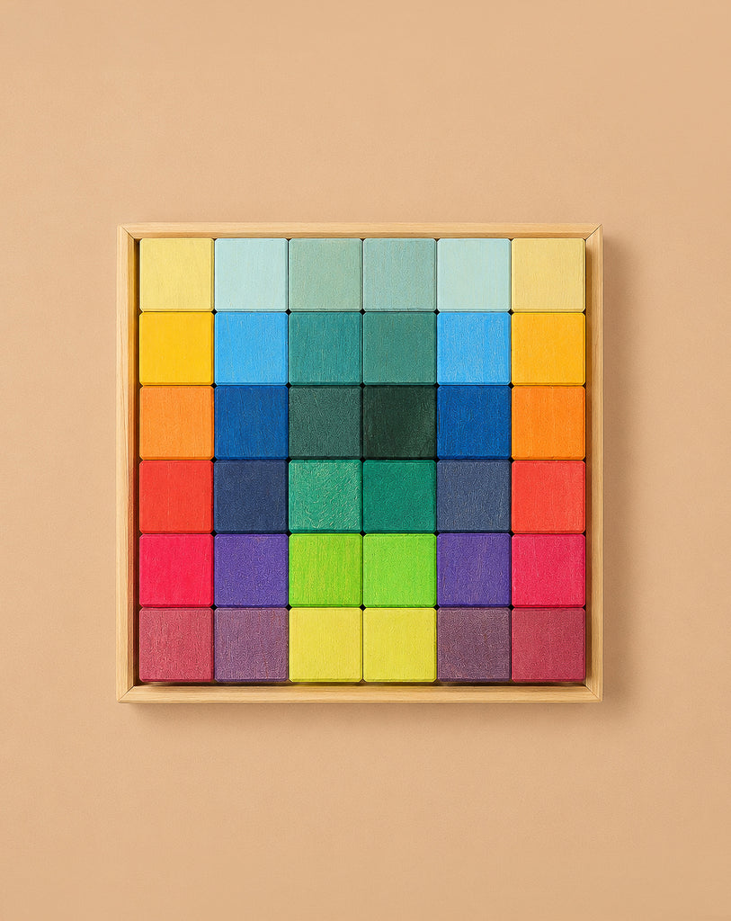 Grimm's Rainbow Mosaic Building Blocks are neatly arranged in a wooden tray, featuring a symmetrical, gradient pattern that transitions from warm reds, oranges, and yellows along the edges to cool greens, blues, and purples at the center—ideal for promoting fine motor skills and spatial thinking through engaging play.