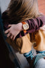 A child with long hair rests their head on folded arms, wearing a burgundy sweater, yellow pants, sneakers, and the Mini Kyomo Watch - Cherry Bomb—a fun kids watch for learning to read time.