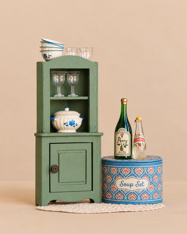 A miniature green Maileg China Cabinet Set holds small bowls, glassware, and a lidded tureen, beside a blue patterned tin topped with toy bottles labeled “Perry” and “Lemonade.”