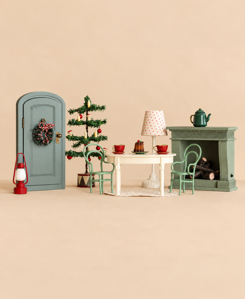 A Maileg Christmas Room Set featuring a door with a wreath, a Christmas tree, a dining table with two chairs, two tea cups, a dessert, and a fireplace with a teapot on the mantle.