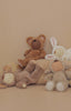 Four plush toys with closed eyes, dressed as a bear, bunny, and two lambs, are arranged on a beige background beside a textured cushion. Spot the cuddly Olli Ella Cozy Dinkum Doll – Lamby Pip among them.