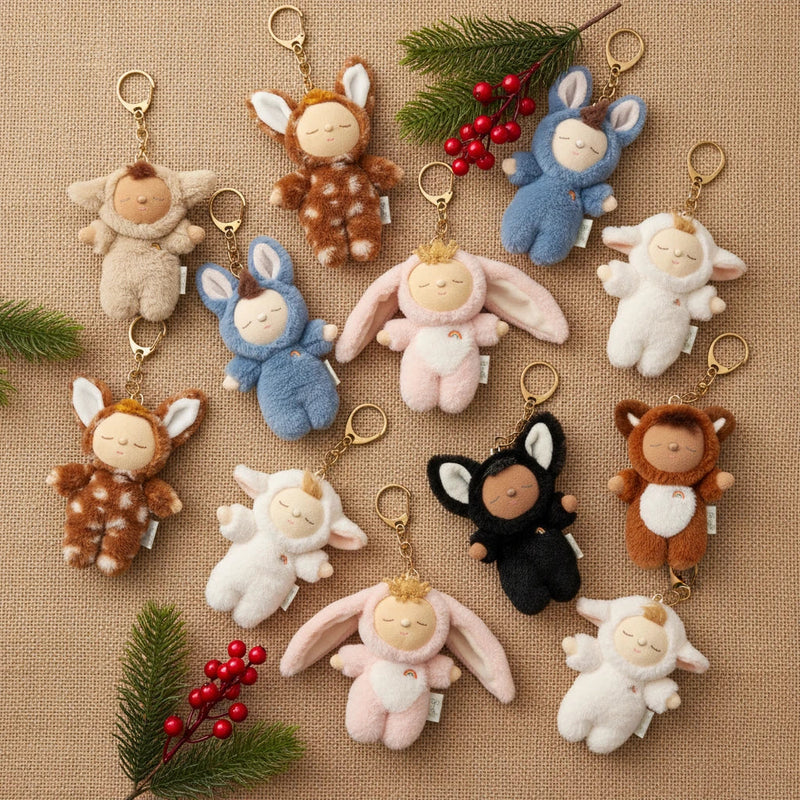 The Olli Ella Cozy Bag Charm - Bunny Twiggy, a plush keychain shaped like a sleeping baby in a bunny outfit, is displayed on burlap with pine branches and red berries.