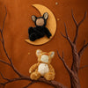 An Olli Ella Cozy Dinkum Doll - Tabby Cat Jinx plush wearing a black cat-ear hood rests on a crescent moon, while a plush orange tabby cat sits on a tree branch below.