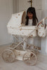 A young girl smiles while playing with the Konges Sloejd Doll Pram - Lemon, a vintage-inspired pram with large wheels and lemon patterns, in a bright room featuring light wooden floors and white walls.