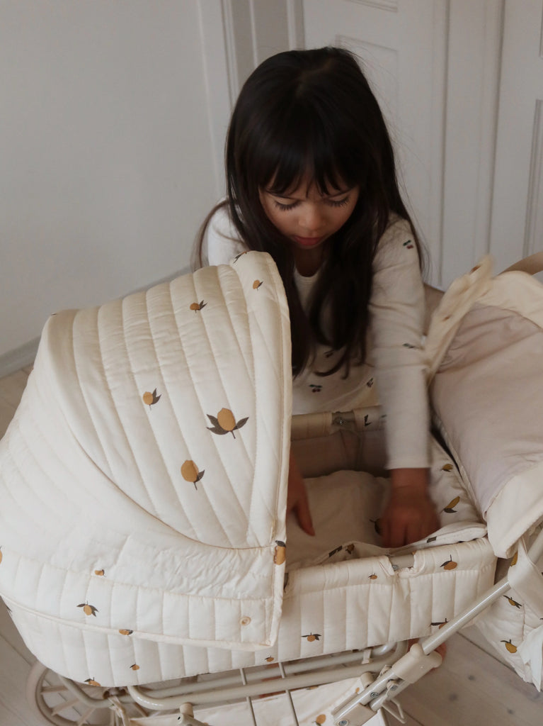 A young girl with long dark hair stands beside the cream-colored Konges Sloejd Doll Pram - Lemon, featuring a citrus fruit pattern and organic cotton mattress, gently placing her hands inside. The room has light walls and a calm mood.