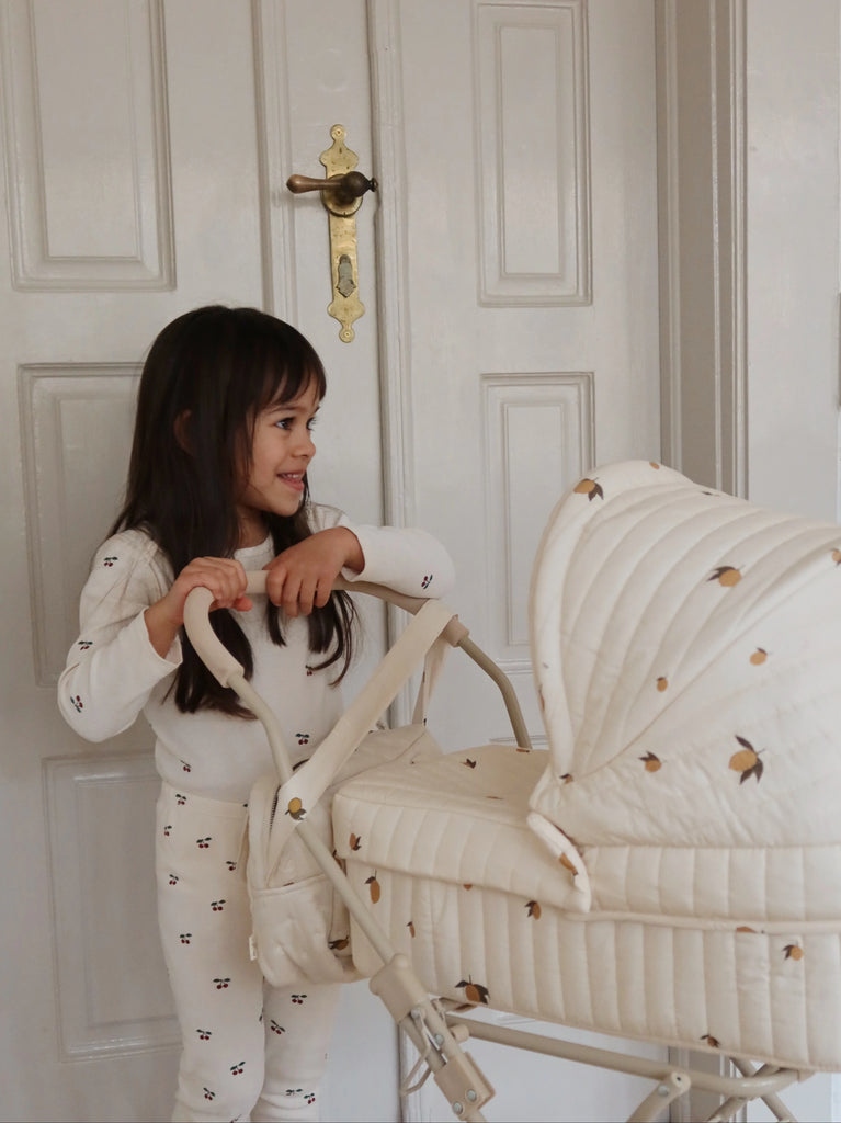 A young girl in white pajamas smiles while holding the handle of the Konges Sloejd Doll Pram - Lemon, featuring large wheels, standing indoors in front of white paneled doors.