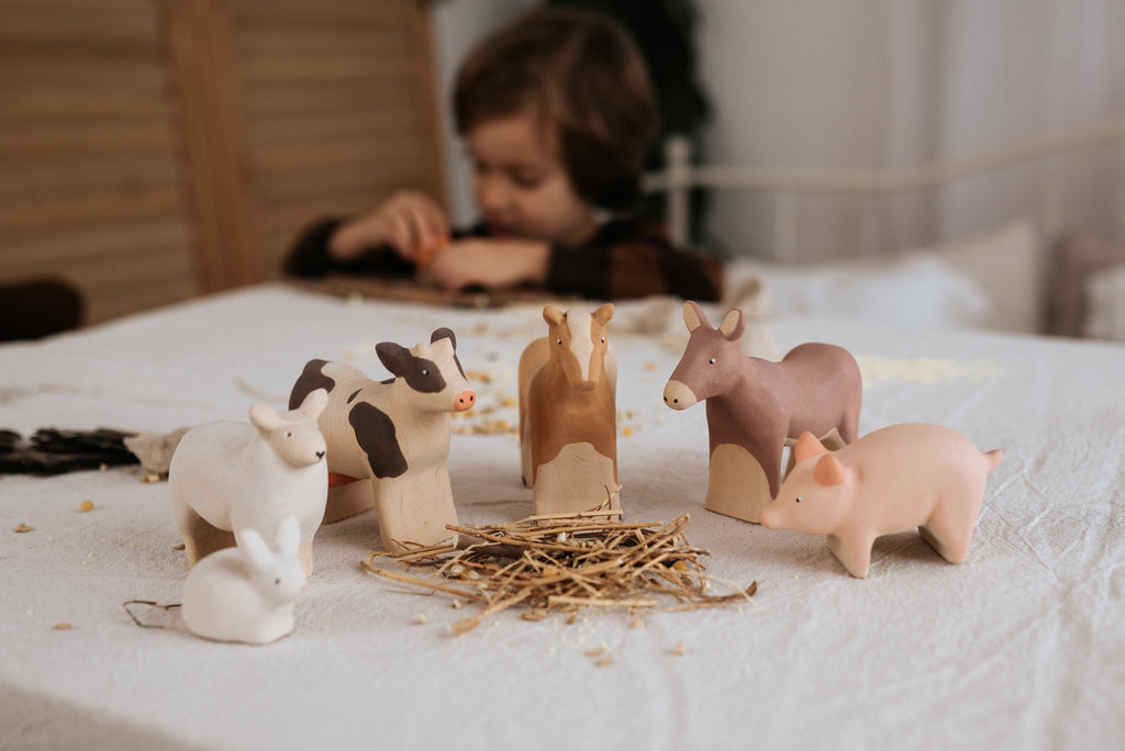 A child plays nearby as handcrafted linden wood farm animal figurines, including a Handmade Wooden Donkey adorned with child-safe paint, are arranged around sticks on a white tablecloth.