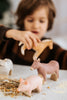 A child enjoys handmade wooden toys—a pink pig, a cow, and a Handmade Wooden Donkey—all crafted from linden wood with child-safe paint. These charming farm animals sit on a hay-covered table as the child plays in the background.